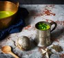 Metal jug and bowl filled with green vegetable soup, next to wooden spoon